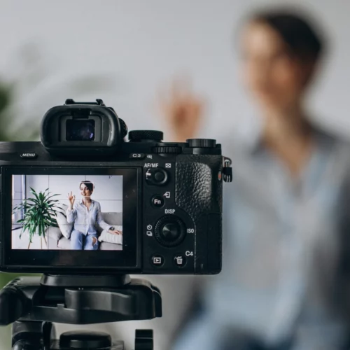 Photo illustrant le service de mise à disposition d'un livret d'accueil vidéo Une caméra enregistre une personne floue en arrière-plan, assise sur un canapé avec une plante verte à côté. L'écran de la caméra montre une image nette de la personne saluant.