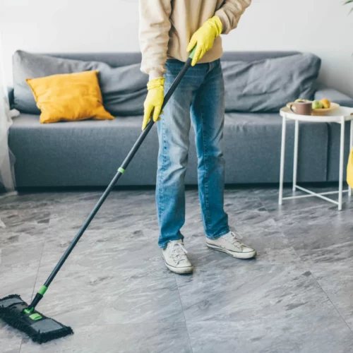 Découvrez les services de conciergerie. Photo illustrant le service de gestion du ménage. Un homme portant des gants jaunes nettoie le sol avec une serpillière dans une pièce bien rangée, comprenant un canapé gris et un fauteuil jaune.